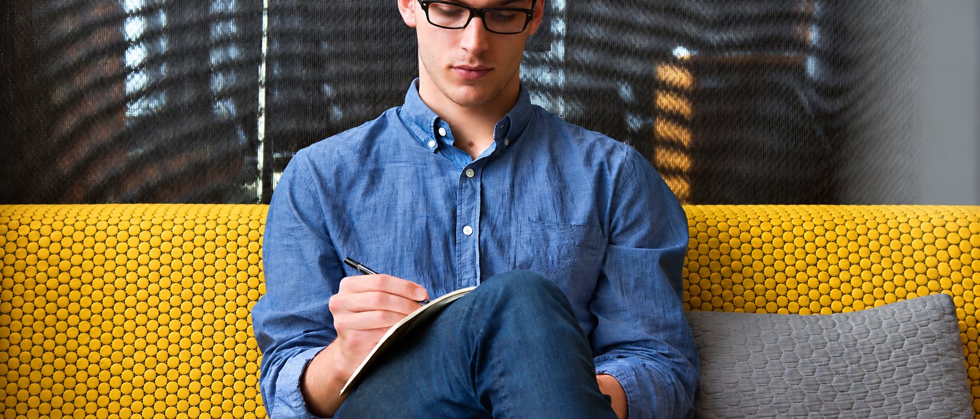 Un jeune homme à lunettes, en chemise bleue, est assis sur un canapé jaune alvéolé et écrit dans un carnet.